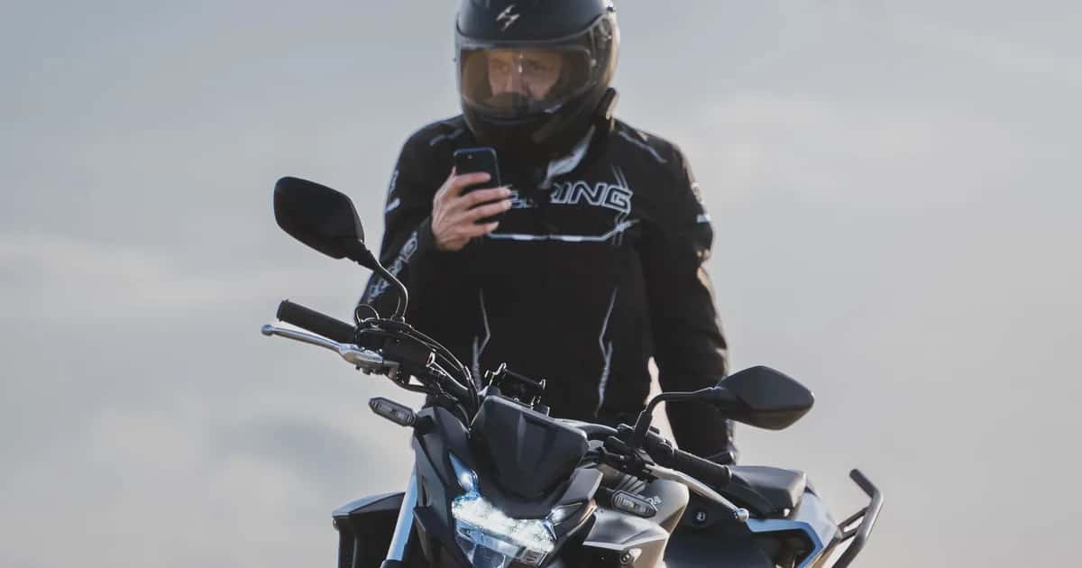 Motociclista de capacete consulta o celular montado na moto, em ambiente aberto