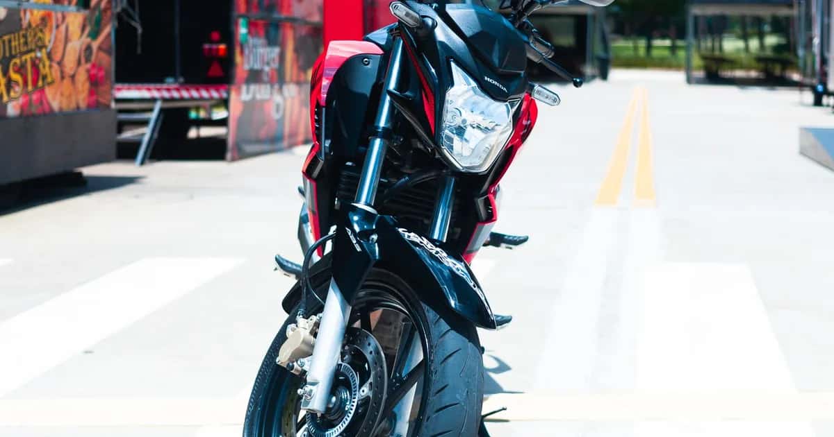 Moto naked Honda de baixa cilindrada estacionada em praça urbana, com prédios ao fundo