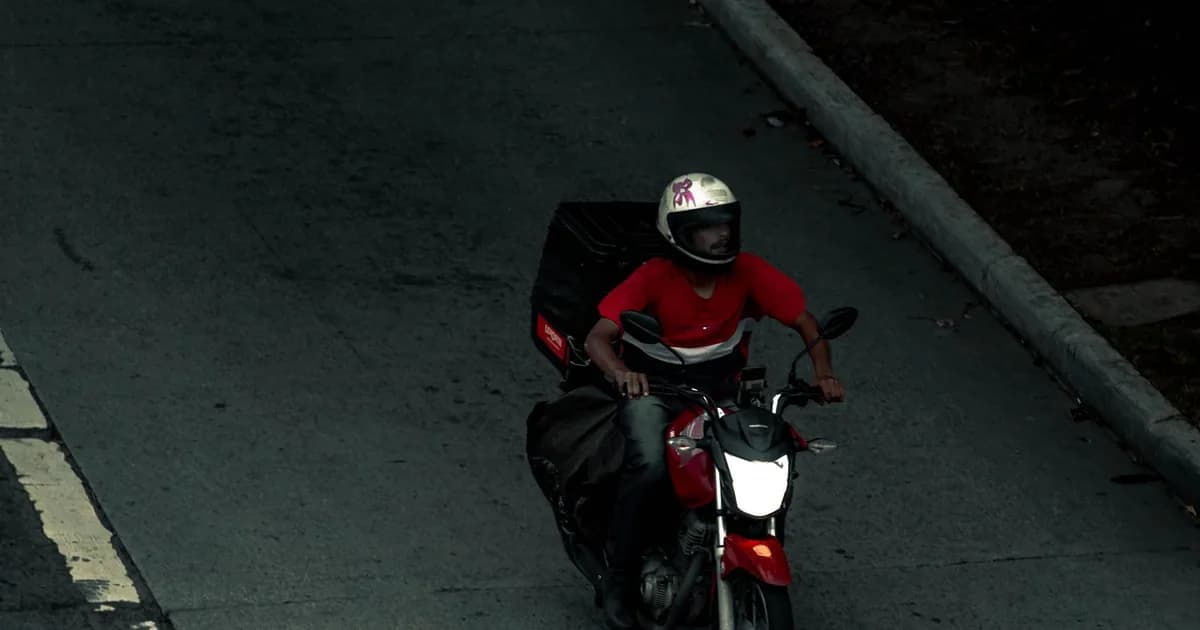 Motoboy de capacete pilota uma moto vermelha de baixa cilindrada com baú de entrega em avenida urbana, visto de cima