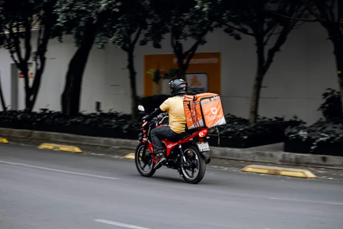 Entregador de aplicativo com baú térmico roda em avenida urbana arborizada