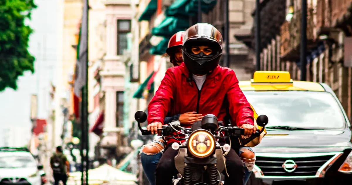 Motociclista de capacete conduz passageira na garupa em rua de centro urbano, com táxi ao fundo