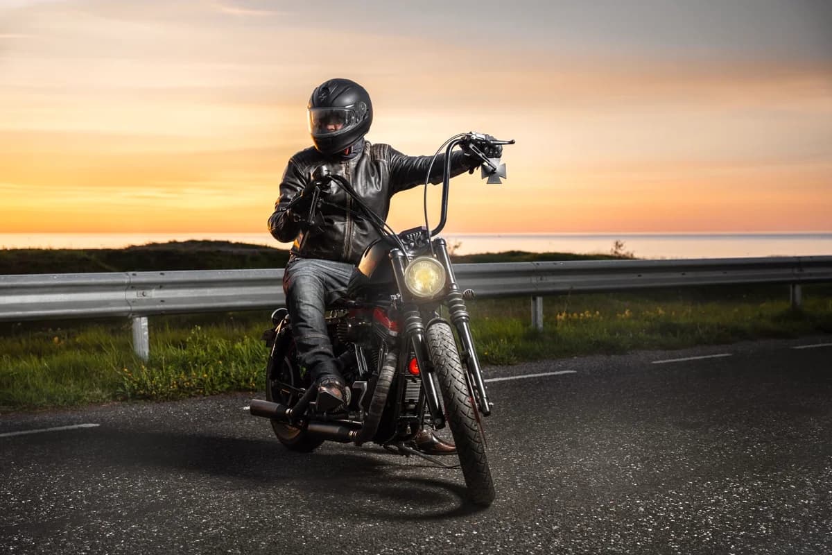 Motociclista de jaqueta de couro em estrada costeira ao pôr do sol