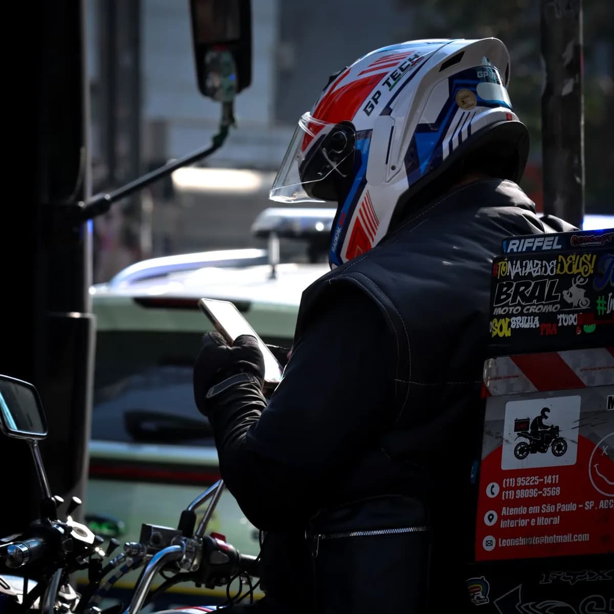Entregador de capacete consulta o aplicativo no celular ao lado da moto, em rua urbana