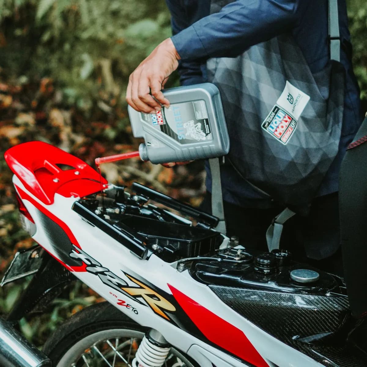 Motociclista de capacete verte óleo no motor de uma moto ao ar livre, com o frasco inclinado sobre o bocal